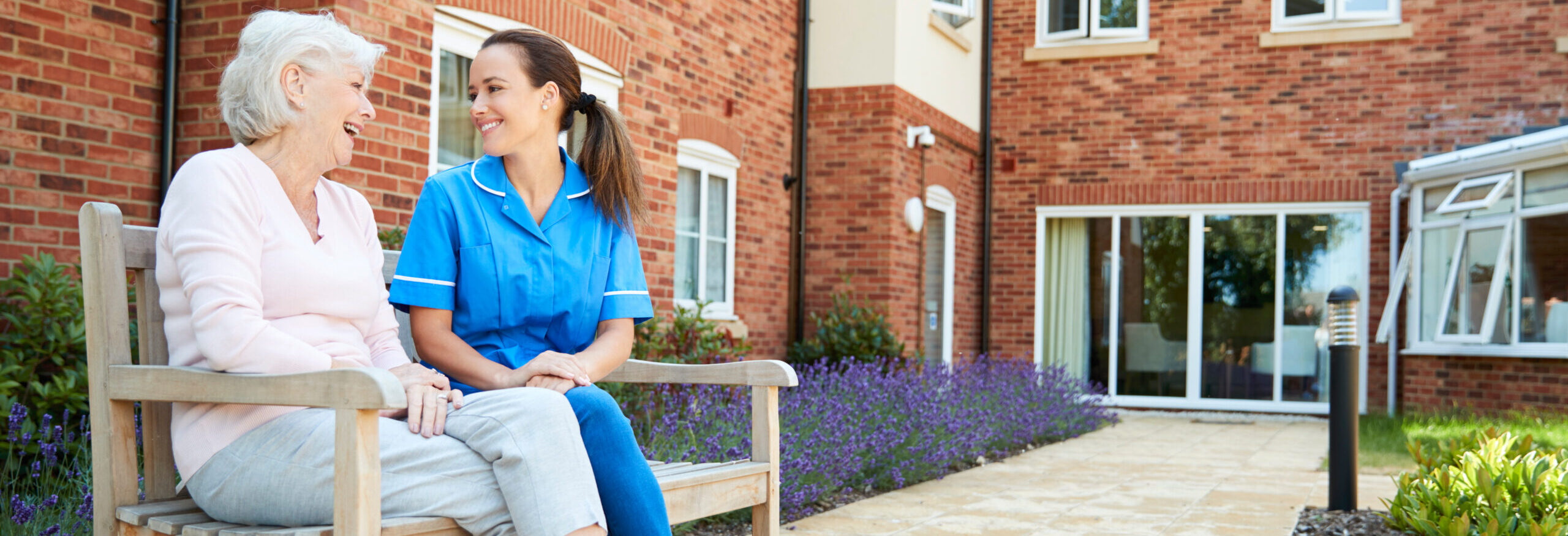 Senior Woman Sitting On Bench And Talking With Nurse In Retirement Home